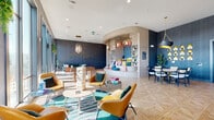 This single-floor amenity space is designed with a bold, contemporary aesthetic that blends playful energy with sophisticated style. The interior is anchored by deep navy-toned textured wall coverings, warm parquet-patterned hardwood flooring, and recessed lighting complemented by eye-catching pendant fixtures in varied shapes and finishes.

The main lounge area features a cluster of cognac leather chairs arranged around low round tables atop a vibrant teal-patterned rug, with a Jenga tower serving as a centerpiece gaming feature. A foosball table occupies the sunlit window-facing zone, where floor-to-ceiling glass doors flood the space with natural light and frame sweeping urban views. A teal velvet sectional and rust-toned lounge chairs create an additional relaxed seating vantage point.

A curved architectural nook with built-in shelving and colorful cushioned seating adds a whimsical social corner, while a separate dining area features an oval marble-top table beneath a dramatic drum pendant with a gold interior. Gold sculptural wall accents and curated greenery punctuate the space throughout.

The covered outdoor terrace offers artificial turf, wire lounge chairs, a projector, and ceiling fans, all surrounded by open railings showcasing expansive city skyline views.