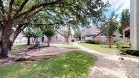 This single-floor residence is part of a classic brick building complex nestled among mature oak trees and manicured green lawns, offering a serene outdoor environment with walkways and communal seating areas. The exterior evokes a well-established community with traditional architectural character.

Inside, the space is finished with smooth, neutral-toned walls that provide a clean, gallery-like backdrop throughout. The flooring consists of polished concrete, lending an industrial-modern aesthetic that flows consistently across the interior. Recessed lighting illuminates the rooms with a warm, focused glow.

The dining area serves as a standout feature, anchored by a substantial round green marble table paired with woven metal chairs atop a natural jute rug. A large framed abstract artwork adorns the wall, adding an artistic focal point. A sleek sliding barn door on a black metal track connects rooms with both style and practicality, contributing to the open, contemporary feel of the layout.

Adjacent spaces are visible through antique-style mirrored doors, hinting at a thoughtfully curated interior design sensibility. The walls display a curated selection of framed art pieces, reinforcing an elevated, design-forward living experience. Overall, the space balances industrial and refined elements to create a sophisticated and comfortable environment.