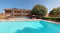 Wilshire Heights offers a welcoming community environment anchored by a generously sized outdoor pool. The pool area is surrounded by a clean concrete deck furnished with multiple lounge chairs, providing ample space for relaxing under open skies. A shaded patio table with umbrella offers an alternative gathering spot, framed by mature trees and well-maintained landscaping. The backdrop features classic two-story brick construction with covered balconies, adding architectural character to the outdoor setting.

The single-floor interior units feature a practical, open layout with neutral-toned carpet throughout the living areas and durable wood-style flooring in connecting spaces. The living room flows directly to a peninsula-style kitchen equipped with dark cabinetry, black appliances including a refrigerator, and a breakfast bar ideal for casual dining. A textured ceiling and soft ambient lighting complete the kitchen area.

Bedrooms are carpeted and finished with lightly textured walls in neutral tones, offering a clean and versatile canvas. Windows fitted with horizontal blinds allow controllable natural light and outdoor views. Hallways connect rooms efficiently, with access panels and utility provisions integrated discreetly into the walls. The overall interior presents a straightforward, functional design suited for comfortable everyday living.