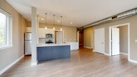 Located at 625 Goodman St S in Rochester, NY, this modern 2-bedroom, 2-bathroom unit offers a well-designed single-floor layout with contemporary finishes throughout. The open-concept living and kitchen area serves as the heart of the home, featuring light wood-look flooring, neutral warm-toned walls, and recessed lighting. A standout feature is the kitchen's bold navy-blue island with a white countertop, complemented by white cabinetry, stainless steel appliances, and sleek pendant lighting overhead. An exposed metallic ductwork element adds an industrial loft-like character to the space. The spacious living area is bathed in natural light from multiple windows and a glass door that frame views of surrounding greenery. Both bedrooms are comfortably sized, with the primary bedroom offering generous proportions, light flooring, and roller shade windows that provide natural light and outdoor views. The second bedroom also features bright windows with tree-lined views. Each of the two bathrooms is finished with large-format tile flooring, dark wood vanities with white countertops, and glass-enclosed walk-in showers. A hallway and dedicated closet provide practical storage. The unit also includes a dedicated entry hallway, tying together a cohesive, move-in-ready living space.
