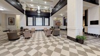 This distinguished lobby showcases a blend of classical architectural elements that create a commanding and refined first impression. The space is defined by soaring double-height ceilings that amplify its grandeur, supported by substantial columns clad in crisp white detailing. A striking checkerboard terrazzo floor in grey and cream tones anchors the space with timeless elegance.

Dark, intricately carved wood frames the entry doors and interior windows, providing a bold contrast against the light-toned walls. Ornate wrought iron railings with brass accents line the stairways, adding sophisticated detailing throughout. Large frosted glass panels set within the dark wood framework allow diffused light to filter into the interior, while an arched window above the secondary entry floods the space with natural light and exterior views.

A prominent clock is mounted above a mezzanine-level balcony, serving as a classic focal point. Black marble wainscoting runs along the lower walls, reinforcing the formal aesthetic. The seating area, furnished with striped and upholstered chairs arranged around low tables, provides a comfortable gathering space. Potted greenery is thoughtfully placed throughout, softening the architectural formality and adding warmth to this stately environment.
