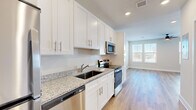 This efficiently designed single-floor unit offers a clean, modern aesthetic throughout, with light wood-tone flooring and crisp white walls creating a bright, cohesive atmosphere. The galley-style kitchen is a standout feature, equipped with white shaker cabinets, granite countertops, a stainless steel undermount sink, and a full suite of stainless steel appliances including a refrigerator, electric range, over-the-range microwave, and dishwasher. Ample upper and lower cabinetry provides generous storage. The living area is bathed in natural light from multiple windows fitted with blinds, and a ceiling fan with an integrated light fixture adds both comfort and function. The neutral wall tones and hardwood-style flooring carry seamlessly throughout, lending a sense of openness. Two bathrooms mirror the unit's consistent design language, featuring granite-topped white vanities, brushed nickel hardware, and a tub-shower combination with tiled surround in one bath. A dedicated closet with louvered doors offers practical storage. The entryway is neatly defined, providing a clean transition into the living spaces. Overall, the unit presents a modern, move-in-ready environment with quality finishes and thoughtful layout.