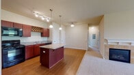 Located at 23728 Springs Ct in Plainfield, IL, this single-floor unit offers a well-organized layout with a seamless flow between the kitchen, living area, and private bedrooms. The kitchen is a standout feature, showcasing rich dark-wood cabinetry, granite countertops, a large center island with pendant lighting above, and a full suite of black appliances including a refrigerator, range, microwave, and dishwasher. A decorative wine rack built into the upper cabinetry adds a distinctive touch, while track lighting and hardwood-style flooring complete the space. The kitchen opens directly into the living area, which transitions to plush carpeting and centers around a tiled fireplace framed by a classic white mantel. A ceiling fan and a window providing natural light enhance the comfort of this area. The bedrooms are carpeted and each equipped with a ceiling fan; one bedroom includes multiple closet doors and connects to a hallway. The bathroom features tile flooring, a granite-topped vanity with dark wood cabinetry, and a full tub and shower combination. Overall, the unit presents a cohesive, functional design with warm finishes throughout.