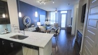 This single-floor residence offers a thoughtfully designed open-concept layout that seamlessly connects the kitchen, dining, and living areas. The kitchen is a standout feature, boasting a generous island topped with light-veined marble countertops, stainless steel appliances including a dishwasher, and pendant lighting that adds a polished, contemporary touch. A deep farmhouse-style sink and clean white cabinetry complete the space with functional elegance.

The dining area flows naturally from the kitchen, providing an inviting setting for everyday meals or entertaining guests. Rich, dark hardwood flooring runs throughout, grounding the space with warmth and visual continuity. The living area is anchored by a bold navy blue accent wall that creates a striking focal point, complemented by tasteful décor including a statement mirror and layered area rug.

Natural light pours in through large windows fitted with sheer curtains, brightening the interior and highlighting the open, airy feel of the space. A ceiling fan with a light fixture enhances comfort and adds a subtle decorative element. Track lighting along the ceiling provides additional illumination and a modern industrial accent. The overall ambiance is contemporary and sophisticated, blending bold design choices with warm, livable details throughout.