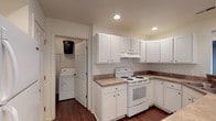 This single-story unit at The Brickyard offers a well-organized, functional layout with a clean, neutral aesthetic throughout. The entry foyer welcomes residents with warm carpet flooring, a chandelier overhead, and a hardwood transition at the front door, creating a polished first impression.

The kitchen is a standout feature, outfitted with white shaker-style cabinetry, dark laminate countertops, and recessed lighting. White appliances include an electric range with an overhead exhaust hood. Directly adjacent to the kitchen is an in-unit laundry area, conveniently tucked behind a paneled door and equipped with a washer and dryer. Rich hardwood-style flooring extends throughout the kitchen and laundry area.

The living area is generously sized and carpeted, featuring a ceiling fan and a sliding glass door with vertical blinds that opens to an outdoor area, allowing natural light to fill the space. A secondary exterior door is also present in the living area.

The bedrooms are carpeted and fitted with ceiling fans. At least one bedroom includes a walk-in closet with built-in hanging storage. The bathroom features hardwood-style flooring, a full tub-shower combination with a curved curtain rod, a toilet, and a white vanity.