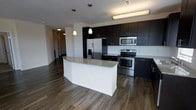 This single-floor space showcases a contemporary open-concept kitchen designed with a clean, modern aesthetic. The kitchen is anchored by a large central island with a white base and a speckled granite countertop, providing ample workspace and a natural gathering point. Dark espresso-toned cabinetry lines the walls from counter height to ceiling, offering generous storage while creating a bold visual contrast against the light-colored walls and backsplash. Stainless steel appliances, including a French door refrigerator with an ice and water dispenser, an electric range with oven, and a built-in microwave, lend a polished, professional quality to the space. Warm hardwood-style flooring runs throughout, adding texture and continuity. Pendant lighting hangs above the island, complementing the flush-mount ceiling fixtures to ensure well-distributed illumination. The open layout connects the kitchen seamlessly to adjacent living areas visible in the background, enhancing the sense of spaciousness. A hallway leading to additional rooms is visible, suggesting a practical and well-organized floor plan throughout the residence.