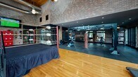 Located on Commerce Drive in Exton, PA, this single-floor fitness facility combines industrial character with comprehensive workout amenities. The space is defined by its striking architectural design, featuring soaring ceiling heights, expansive floor-to-ceiling steel-framed windows, exposed brick walls, warm wood-plank ceilings, and polished hardwood floors throughout. Visible ductwork and pendant lighting reinforce the industrial aesthetic while maintaining a welcoming atmosphere.

The main workout floor is generously proportioned and anchored by a full-size boxing ring, complemented by a dedicated strength and combat zone housing heavy bags, free weights, and functional training equipment. Rows of cardio machines—including treadmills, ellipticals, stationary bikes, and a stair climber—are positioned along the expansive window walls, providing ample natural light and views of the surrounding area.

A separate studio space features frosted glass partition walls, polished hardwood floors, a wall-mounted display screen, and a ballet barre, making it well-suited for group fitness classes, yoga, or movement training. The open layout throughout the facility supports a variety of training disciplines, and the abundant natural light flowing in from multiple window walls creates an energizing environment conducive to focused physical activity.
