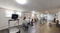 Located at 2920 Dean Parkway in Minneapolis, Minnesota, this fitness center offers a single, well-utilized exercise room designed to support a comprehensive workout experience. The space features durable rubber flooring throughout, providing both safety and comfort during exercise. Cardio equipment includes treadmills, an elliptical machine, and stationary bikes, catering to a range of aerobic workout preferences. The strength training area is equipped with a barbell rack, weight bench, and a full dumbbell rack, accommodating users of varying fitness levels. Stability balls and additional accessories round out the functional training options. A large wall-mounted mirror on one end of the room enhances the sense of space and allows users to monitor their form, while a wall-mounted flat-screen television adds entertainment during workouts. Informational fitness posters are displayed along the white walls, offering exercise guidance. The room is finished with a textured ceiling and flush-mount ceiling lights that provide consistent, even illumination throughout. A wall-mounted air conditioning unit ensures comfort year-round. A security camera is also present, contributing to a safe environment. The space is accessed through a single door, making it a self-contained, functional fitness facility.