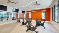 This single-level spin studio is thoughtfully designed to deliver an energizing workout environment. The space is anchored by a fleet of stationary cycling bikes arranged across light-toned hardwood flooring, offering ample room for structured cycling classes. Bold orange accent walls serve as a defining visual element, creating a high-energy atmosphere that complements the room's active purpose.

The ceiling is a standout architectural feature, showcasing sweeping curved LED light strips that trace fluid, wave-like patterns across the white surface. Ceiling fans are strategically positioned throughout to promote airflow during intense sessions. Full-length mirrors line one wall, enhancing the sense of depth while allowing participants to monitor their form.

Wall-mounted flat-screen televisions provide entertainment or instructional content during classes. Large black-framed windows along multiple walls flood the studio with natural light and offer views of the surrounding outdoor environment. Near the entrance, a white accent wall features an artistic orange line-work mural, adding a creative design touch that ties into the room's color palette. The overall ambiance is modern, vibrant, and purpose-built for high-performance group fitness.