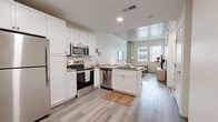 This thoughtfully designed one-bedroom, one-bathroom residence offers a cohesive blend of modern industrial aesthetics and contemporary comfort across a single floor. The open-plan kitchen and living area forms the heart of the home, where striking exposed concrete ceilings add an urban, loft-inspired character. The kitchen is outfitted with white shaker cabinetry, stainless steel appliances including a refrigerator, range, microwave, and dishwasher, and a peninsula with a grey quartz countertop that provides both additional workspace and casual bar seating. Light wood-look flooring runs throughout the main living areas, complementing the neutral, bright palette. The living room benefits from large windows that invite generous natural light and urban views, creating an airy and inviting atmosphere accentuated by an exposed concrete column detail. The bedroom is a calm retreat finished with soft carpeting, a large window, and the same soaring concrete ceiling feature. A dedicated closet provides practical storage. The bathroom is clean and contemporary, featuring a glass-enclosed shower, a white vanity with a grey countertop, a framed mirror, and matching light wood-look flooring. Recessed lighting throughout ensures a well-lit environment in every room, making this a highly functional and stylish urban residence.