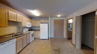 This single-floor unit combines a kitchen and living area in a practical, open layout. The kitchen is outfitted with light wood cabinetry providing ample storage across both upper and lower cabinets. White appliances — including a refrigerator, electric range, and over-the-range microwave — are included. Dark laminate countertops offer a subtle contrast to the cabinetry, and a sink with a window above it adds natural light to the cooking area. The flooring transitions from a tile pattern in the kitchen to carpet in the adjacent living space, clearly delineating the two functional zones. Flush-mount ceiling fixtures provide overhead lighting throughout. A solid entry door leads directly into the main area, offering a convenient and straightforward layout. Just off the main space, a bathroom is visible through an open doorway, featuring a vanity with a mirror and lighting above. The neutral color palette — gray walls, light wood tones, and white appliances — gives the space a clean, understated aesthetic suited for everyday living.