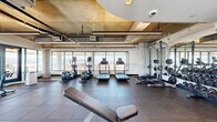 This fitness center in Denver, CO offers a generously sized, single-floor layout comprising a main exercise room and a connecting hallway. The space is defined by its industrial-modern aesthetic, featuring exposed concrete ceilings with visible ductwork and piping that lend an urban, loft-like character to the environment.

Floor-to-ceiling windows wrap around much of the exercise room, flooding the space with abundant natural light and providing sweeping panoramic views of the Denver skyline and the surrounding Rocky Mountain landscape. The rubber tile flooring throughout the workout area provides both durability and safety underfoot.

The exercise room is comprehensively equipped to support a wide range of fitness activities. Cardio equipment includes treadmills, elliptical trainers, stationary spin bikes, and a rowing machine. Strength training is well-represented with a squat rack, a full dumbbell rack, a cable machine, and adjustable benches. Stability balls and functional training accessories round out the offerings, making the space suitable for varied workout routines.

The hallway entry area features clean white walls, recessed lighting, and a large interior window that maintains visual connectivity to the main fitness floor. Together, the rooms form a cohesive, thoughtfully designed fitness facility that balances functionality with an inspiring, light-filled atmosphere.
