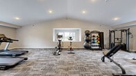 This fitness center offers a generous, open workout environment thoughtfully designed to accommodate a wide variety of exercise routines. The space is defined by its striking vaulted ceiling, which rises to create an airy, expansive atmosphere enhanced by recessed lighting throughout. Neutral-toned walls and patterned carpet tile flooring give the space a clean, modern aesthetic.

The facility is organized to support both cardio and strength training. The cardio section houses treadmills, stationary spin bikes, an elliptical machine, and a rowing machine, providing ample options for aerobic exercise. Multiple windows bring in natural light and offer views of the surrounding greenery, creating an inviting environment for workouts. Full-length wall mirrors reflect the space and assist with form during exercise.

The strength training area is well-appointed, featuring a multi-function cable machine, a comprehensive dumbbell rack with a full range of weights, a medicine ball and stability ball storage rack, and an adjustable workout bench. Storage closets are integrated into the layout, helping keep the space organized. An accessible entry door with an emergency exit sign ensures safety and convenience. Overall, this fitness center delivers a complete and functional workout facility in a well-maintained, comfortable setting.
