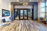Located at 119 Searing Avenue in Mineola, NY, this single-floor commercial space presents a polished and contemporary lobby environment designed with both functionality and aesthetic appeal in mind. The entrance is defined by dramatic variegated wood-look flooring in cool grey and natural tones, which flows seamlessly throughout the space and establishes an immediate sense of sophistication.

The lobby features bold architectural elements, including dark-framed glass double doors etched with signage, flanked by contrasting textured white and deep charcoal accent walls. A cascading pendant chandelier with exposed Edison-style bulbs adds an industrial-modern focal point overhead, complementing the recessed lighting throughout the space.

A dedicated workstation area near the reception zone is anchored by sleek modern furnishings and framed art that adds a curated, professional character. Striped transitional carpeting demarcates this functional zone with subtle elegance.

An elevator and a staircase with dark-trimmed steps and a clean metal railing provide vertical access within the building. The lounge area is highlighted by oversized floor-to-ceiling grid windows that invite abundant natural light and offer street-level views of the surrounding Mineola neighborhood. A low-profile leather sofa, a sculptural blue glass coffee table, and warm drapery complete this welcoming seating vignette, making the overall space feel both professional and inviting.