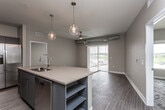 This single-floor unit showcases a thoughtfully designed open-concept layout that seamlessly connects the kitchen and living area. The kitchen is a standout feature, equipped with stainless steel appliances including a refrigerator, range, and microwave, complemented by sleek gray cabinetry, warm butcher block-style countertops, and a striking chevron-patterned tile backsplash. A large central island with a built-in sink and pendant globe lighting serves as both a functional workspace and a natural gathering point. Exposed ductwork running along the ceiling adds an industrial character that distinguishes the space.

The living area is bright and open, benefiting from a sliding glass door that invites natural light and provides outdoor access. A ceiling fan enhances comfort, while wood-look flooring flows consistently through the main living spaces. The bedrooms feature plush patterned carpeting and generous walk-in closets outfitted with wire shelving systems for ample organization.

Two bathrooms serve the unit: one with a dual-sink vanity with gray cabinetry and a separate walk-in shower with decorative tile, and a second with a full tub-shower combination. The neutral warm-toned walls and clean white trim throughout create a cohesive, contemporary aesthetic that is both functional and visually inviting.