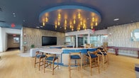 Located at 205 Revere Beach Parkway in Revere, Massachusetts, this single-floor amenity lounge is a thoughtfully designed social space that blends modern aesthetics with comfort and functionality. The centerpiece of the room is a striking curved bar finished in bold blue, topped with a sleek white countertop and surrounded by wooden bar stools with blue upholstered seats. Above the bar, a dramatic circular ceiling medallion houses an array of amber pendant lights that cast a warm, inviting glow throughout the space. The lounge area features generous seating, including plush sofas and eclectic accent chairs arranged around a large flat-screen television mounted against a textured wood-paneled wall. A standout fireplace framed in veined stone tile serves as an elegant focal point in a separate seating alcove, with wall sconces and sheer curtains adding a refined touch. The kitchen area is equipped with under-counter refrigeration units and a large island, making the space well-suited for entertaining. Floor-to-ceiling windows along one side of the space flood the interior with natural light while offering views of the surrounding neighborhood. Light hardwood flooring, a dark slat ceiling, and glass-walled private rooms complete the polished, contemporary design of this versatile community amenity space.