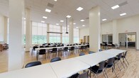 This expansive single-floor facility is designed to accommodate a wide range of gatherings and activities. The main hall is defined by its notably high ceilings and a grid of structural columns finished in a neutral cream tone, contributing to an open and airy atmosphere. Large floor-to-ceiling windows line the perimeter walls, fitted with roller shades to allow precise light control, while providing broad views of surrounding greenery and landscaped outdoor areas.

The flooring throughout is light-toned hardwood, lending warmth to the otherwise clean, modern interior. Recessed and flush panel ceiling lights ensure consistent, even illumination across the space. Multiple rows of long white folding tables paired with black stackable chairs are arranged throughout the hall, offering a highly flexible seating configuration suitable for lectures, dining, meetings, or community events.

A ceiling-mounted projector and a wall-mounted flat-screen television provide built-in audiovisual capability. A retractable projection screen is also present, further supporting presentations or screenings. A smaller adjoining room features an upright piano and a round table, offering a quieter supplementary space. The overall design is functional and contemporary, prioritizing versatility and natural light.