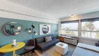 Located in the heart of Downtown Los Angeles, this thoughtfully designed studio at 725 S Bixel St offers a compelling blend of contemporary style and practical living. The open-plan layout seamlessly integrates the sleeping, living, and kitchen areas into a cohesive, well-appointed space.

The living area is anchored by a bold teal accent wall, which adds depth and character to the room. A comfortable sofa, a compact dining nook with a yellow table, and a multi-directional floor lamp contribute to the space's modern, curated aesthetic. A large window fills the room with natural light and frames views of the surrounding cityscape and palm trees.

The sleeping area features a platform bed with built-in storage and a wall-mounted flat-screen TV, positioned against the same striking teal wall. A built-in desk sits adjacent to the bed, maximizing functionality within the studio footprint.

The kitchenette is clean and efficient, outfitted with dark cabinetry, a white countertop, an undermount sink with a matte black faucet, and open shelving. Polished concrete floors run throughout, lending an industrial-chic quality to the overall design.

The bathroom maintains the space's crisp aesthetic with a wide vanity, a large mirror with overhead lighting, classic white tile flooring, and a shower-tub combination with a bold black curtain.