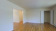 This single-floor space offers a straightforward and functional layout characterized by its warm, honey-toned hardwood floors that run continuously throughout, lending the interior a cohesive and inviting feel. The walls are finished in a crisp, neutral off-white tone, creating a bright and airy atmosphere that allows for versatile decorating possibilities. The main living area is generously proportioned, providing ample room for a variety of furniture arrangements. A wide, white-trimmed doorway connects the main living space to an adjacent room, suggesting an open and accessible flow between areas. The clean white baseboards and door trim throughout provide a classic finishing touch that complements the simplicity of the overall design. A paneled white door on the far wall leads to an additional space, further extending the unit's connectivity. The ceilings are smooth and of standard height, contributing to the room's clean lines. The overall ambiance is calm, light-filled, and move-in ready, with the natural wood flooring serving as the defining design element of the space.