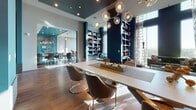 This single-floor space is a thoughtfully designed communal lounge that blends modern aesthetics with practical functionality. The interior is anchored by a generous rectangular white dining or gathering table surrounded by sleek, upholstered chairs in warm taupe tones. Overhead, a sculptural chandelier composed of globe-shaped glass bulbs on brass arms serves as a striking focal point, casting ambient light across the room.

Deep teal accent walls provide a bold backdrop throughout the space, complemented by warm wood flooring laid in a distinctive herringbone pattern. A floor-to-ceiling built-in bookcase, finished in navy blue with integrated lighting, lines one wall and is curated with books, decorative objects, and art pieces. A low upholstered bench in front of the shelving invites leisurely browsing.

A large-scale abstract painting in vivid blues, yellows, and oranges commands attention on a textured stone-clad accent wall, adding an artistic energy to the room. Sheer patterned drapery frames tall windows that flood the space with natural light and offer views of the surrounding landscape.

An adjacent glass-walled conference or co-working room is visible from the main lounge, providing a semi-private workspace option. Together, these areas create a versatile, design-forward environment suited for both social gatherings and productive work.