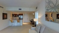 This single-floor space is designed with an open-concept layout that seamlessly connects the kitchen, dining, and living areas, creating a functional and cohesive environment. The kitchen is outfitted with dark wood cabinetry, stainless steel appliances including a refrigerator and microwave, and a light-toned countertop that extends into a breakfast bar with bar-height seating. A undermount sink and clean backsplash contribute to the kitchen's polished appearance.

Adjacent to the kitchen, a compact dining area accommodates a rectangular table with upholstered chairs, illuminated by a ceiling-mounted light fixture that provides warm, focused lighting over the space. The transition into the living area is natural and unobstructed, where a cushioned sofa with decorative throw pillows anchors the room. A table lamp with a classic white shade adds layered lighting and a refined touch.

Throughout the space, light-toned flooring and neutral wall colors create a bright, airy atmosphere. The layout is practical and well-proportioned, with each zone clearly defined yet visually connected, making the overall space feel open, welcoming, and move-in ready.