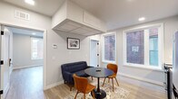 This single-floor unit showcases a clean, modern design with light hardwood-style flooring and crisp white walls that flow consistently throughout the space. The open floor plan, visible in the 3D layout, arranges the kitchen, living area, bedroom, and bathroom in a compact yet functional configuration.

The living area is anchored by large windows that frame views of neighboring brick architecture, filling the room with natural light. A wall-mounted television, framed wall art, and a coffered ceiling detail above the seating area add character and visual interest. The space comfortably accommodates a small sofa and a round dining table with chairs.

The kitchen is well-appointed with two-tone cabinetry — white upper cabinets and grey lower units — complemented by a white tile backsplash, white countertops, and stainless steel appliances including a range, microwave, dishwasher, and refrigerator. Under-cabinet lighting adds a polished finishing touch.

A separate bedroom benefits from a tall, narrow window offering urban views of the surrounding brick buildings, providing natural light while maintaining privacy.

The bathroom features a large walk-in shower enclosed by a sliding glass door with sleek matte black hardware, paired with a light wood vanity and dark tile flooring for a contemporary contrast.