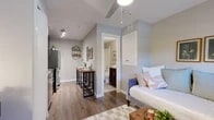 This thoughtfully designed studio unit makes excellent use of its compact single-floor footprint, offering a cohesive and functional living environment. The open-concept kitchen and living area serves as the heart of the space, featuring a fully equipped kitchen with dark wood cabinetry, stainless steel appliances including a refrigerator, electric range, microwave, and dishwasher, and a classic white subway tile backsplash. A sleek bar-height dining table with leather-top stools provides a casual dining area that flows naturally into the living space. Light wood-look flooring runs throughout, lending warmth and visual continuity across the unit. The sleeping area is integrated within the main living space, benefiting from a window with blinds that allows natural light to filter in. A wall-mounted HVAC unit ensures year-round comfort. Recessed lighting and a track lighting fixture brighten the kitchen and living zones effectively. The hallway connects to a full bathroom, which includes a tub-shower combination, a toilet, and a vanity with a granite-style countertop and open wood shelving beneath. Neutral tones and clean finishes throughout give the studio a modern, move-in-ready appeal.