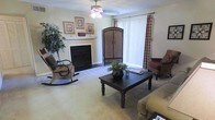 This single-floor residence offers a comfortable and thoughtfully arranged living space with a welcoming, traditional character. The living room serves as the heart of the home, anchored by a classic white-mantled fireplace that adds warmth and a timeless focal point to the room. A ceiling fan overhead provides both function and a subtle decorative touch, while large windows dressed with vertical blinds and plaid curtain panels allow an abundance of natural light to fill the space.

The living room is generously sized, accommodating a mix of seating arrangements including a cushioned rocking chair and accent chairs that contribute to the room's relaxed, homey atmosphere. A dark wood coffee table with turned legs sits at the center, grounded by light-toned carpeting that runs throughout. An ornate armoire adds a bold, traditional design element near the window, complementing the framed artwork displayed on the fireplace mantel.

Neutral wall tones keep the space feeling open and airy, while the warm furnishings and decorative greenery introduce natural textures and color. Overall, the space conveys a cozy, lived-in charm well-suited to comfortable everyday living.
