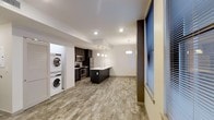 This well-appointed 1-bedroom, 1-bathroom residence is designed with a modern aesthetic and an efficient, open-concept floor plan. The kitchen serves as a central highlight, featuring dark cabinetry, stainless steel appliances including a refrigerator, range, and microwave, as well as a tasteful tile backsplash. A sleek kitchen island with a built-in sink provides additional prep space and casual seating opportunities, while pendant lighting above adds a contemporary touch. The open living area extends seamlessly from the kitchen, finished with durable wood-look flooring that runs throughout, lending warmth and visual continuity to the space. Recessed lighting complements the pendant fixtures, ensuring the interior is well-lit throughout the day. Large windows fitted with blinds allow for natural light while offering privacy control. A standout convenience of this unit is the dedicated in-unit laundry area, which houses a stacked washer and dryer — a practical and sought-after feature. The clean, neutral color palette of white walls and warm flooring creates a bright, inviting atmosphere that is both functional and easy to personalize. The single-floor layout ensures accessibility and ease of movement throughout the entire space.