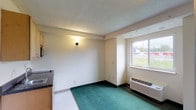 This single-floor studio unit offers a straightforward and functional layout designed to maximize efficient use of space. The main living area features teal carpeting and neutral beige walls, creating a simple and clean environment. A wall-mounted sconce provides ambient lighting, while a window with an exterior view allows natural light to brighten the room. A through-wall HVAC unit ensures climate control year-round.

The kitchenette is well-equipped for everyday cooking and meal preparation, featuring light wood cabinetry — both upper and lower — paired with dark granite-style laminate countertops. It includes a single-basin stainless steel sink, a smooth-top electric cooktop, an under-counter refrigerator, a built-in microwave, and ample cabinet storage above. The transition from carpeted living space to a tile-like entry area near the kitchenette helps define the functional zones within the open floor plan.

The bathroom is clean and utilitarian, finished with speckled vinyl flooring and white fixtures throughout. It includes a full bathtub with overhead shower, a toilet, and a wall-mounted sink with a mirrored medicine cabinet. The textured ceiling throughout the unit adds a subtle architectural detail that ties the space together cohesively.