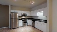 This single-floor space presents a clean, contemporary design that prioritizes both functionality and style. The kitchen serves as a standout feature, outfitted with a full suite of stainless steel appliances, including a refrigerator, gas range, built-in microwave, and dishwasher. Crisp white cabinetry lines the walls from counter to ceiling, offering generous storage while contributing to the bright, open feel of the space. Dark countertops provide a striking contrast against the white cabinets, creating a modern aesthetic that is both visually appealing and practical. The sink is conveniently positioned along the outer wall, complemented by additional counter space ideal for food preparation. Underfoot, light hardwood flooring runs throughout, adding warmth and continuity to the overall design. The kitchen is illuminated by track lighting mounted to the ceiling, ensuring even, adjustable light across the workspace. The layout is well-organized in an L-shaped configuration, maximizing efficiency and ease of movement. The overall ambiance is modern and inviting, with neutral tones and quality finishes that create a polished, move-in-ready atmosphere throughout the space.