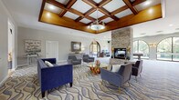 This sophisticated single-floor community clubhouse offers an inviting and elegantly appointed gathering space designed for both relaxation and recreation. The centerpiece of the main lounge is a striking coffered ceiling with rich, dark-stained wood beams and a decorative chandelier that anchors the room with warmth and architectural character. Soaring ceiling heights enhance the sense of openness throughout. The lounge is furnished with plush navy blue sofas and contemporary gray accent chairs arranged around a glass-top coffee table atop a flowing wave-patterned carpet, creating a comfortable and visually dynamic seating area. A floor-to-ceiling stacked stone fireplace adds a dramatic focal point along one wall.

Adjacent to the lounge, an arched doorway leads into a game and activity room featuring a full-sized billiards table, a computer workstation, and a series of large arched windows that frame lush, tree-lined outdoor views. Natural light pours generously throughout the space. A covered patio area with exposed brick walls, hardwood-style flooring, and casual seating offers a relaxed outdoor retreat with views of the surrounding greenery. Arched architectural details are carried consistently throughout the space, lending a cohesive and refined aesthetic. The overall ambiance is polished yet approachable, ideal for community gatherings and everyday leisure.