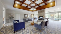 This sophisticated single-floor community clubhouse offers an inviting and elegantly appointed gathering space designed for both relaxation and recreation. The centerpiece of the main lounge is a striking coffered ceiling with rich, dark-stained wood beams and a decorative chandelier that anchors the room with warmth and architectural character. Soaring ceiling heights enhance the sense of openness throughout. The lounge is furnished with plush navy blue sofas and contemporary gray accent chairs arranged around a glass-top coffee table atop a flowing wave-patterned carpet, creating a comfortable and visually dynamic seating area. A floor-to-ceiling stacked stone fireplace adds a dramatic focal point along one wall.

Adjacent to the lounge, an arched doorway leads into a game and activity room featuring a full-sized billiards table, a computer workstation, and a series of large arched windows that frame lush, tree-lined outdoor views. Natural light pours generously throughout the space. A covered patio area with exposed brick walls, hardwood-style flooring, and casual seating offers a relaxed outdoor retreat with views of the surrounding greenery. Arched architectural details are carried consistently throughout the space, lending a cohesive and refined aesthetic. The overall ambiance is polished yet approachable, ideal for community gatherings and everyday leisure.