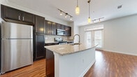This single-floor residence offers a thoughtfully designed open-concept layout where the kitchen flows seamlessly into the living area. The kitchen is a standout feature, showcasing rich dark espresso cabinetry, a mosaic tile backsplash, stainless steel appliances including a refrigerator, range, and microwave, and a generous granite-topped island with a built-in sink and pendant lighting overhead. Track lighting throughout the kitchen adds both function and style, while crown molding lends an elegant finishing touch.

The living area is bright and inviting, featuring warm hardwood flooring, a ceiling fan, and large windows with blinds that allow natural light to fill the space. French-style doors lead to an outdoor area, enhancing the open and airy feel of the room.

The bedroom is carpeted for comfort and includes a ceiling fan and a dedicated walk-in closet with built-in shelving and hanging rods. A second carpeted bedroom also provides walk-in closet access. The bathroom is finished with large-format tile flooring, dark cabinetry with a granite countertop vanity, and a tiled tub-shower combination. Throughout the space, consistent finishes and quality materials create a cohesive and functional living environment.