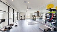 This single-floor fitness studio is thoughtfully designed to support a wide range of exercise disciplines in a bright, open environment. The space is defined by expansive floor-to-ceiling black-framed windows that line the walls, flooding the interior with abundant natural light and providing views of the surrounding exterior. The light gray tile and hardwood-style flooring throughout contributes to the studio's clean, contemporary aesthetic.

Ceiling fans are strategically positioned overhead to ensure comfortable airflow during workouts, complemented by recessed lighting for consistent illumination. Full-length mirrors span a significant portion of one wall, visually expanding the space and allowing users to monitor their form during exercise.

The studio is outfitted with stationary cycling bikes equipped with individual screens, yoga mats laid out in organized rows, stability balls, medicine balls, resistance bands, and a rowing machine — making it well-suited for cycling, yoga, stretching, and functional training. A dedicated equipment storage rack keeps accessories organized and accessible.

Along one wall, built-in cabinetry and open shelving offer additional storage, lending a polished and organized look to the space. A flat-screen display mounted within the mirror wall supports on-demand or instructor-led workout programming. The overall ambiance is clean, airy, and motivating — a professional-grade facility designed to encourage an active lifestyle.