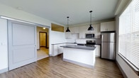 This single-floor space showcases a thoughtfully designed open-concept layout, with the kitchen serving as a central focal point. The kitchen is outfitted with crisp white cabinetry that extends to the upper walls, providing ample storage, while dark countertops on both the perimeter and the central island offer a striking contrast. Two matte black industrial-style pendant lights hang above the island, adding a modern, utilitarian aesthetic to the space.

Stainless steel appliances, including a refrigerator, microwave, and range, are seamlessly integrated into the kitchen design, contributing to a clean and contemporary look. The kitchen island doubles as a functional workspace and casual gathering area. A single-basin sink is positioned along the perimeter counter, complemented by a tile backsplash.

The flooring throughout is a warm-toned wood-look material that anchors the open layout and flows naturally into the adjoining areas. A sliding barn-style door is visible along one wall, adding a rustic design element and providing access to an adjacent room. The overall palette of whites, grays, and natural wood tones creates a bright, cohesive, and inviting atmosphere throughout the space.