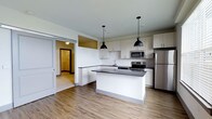 This single-floor space showcases a thoughtfully designed open-concept layout, with the kitchen serving as a central focal point. The kitchen is outfitted with crisp white cabinetry that extends to the upper walls, providing ample storage, while dark countertops on both the perimeter and the central island offer a striking contrast. Two matte black industrial-style pendant lights hang above the island, adding a modern, utilitarian aesthetic to the space.

Stainless steel appliances, including a refrigerator, microwave, and range, are seamlessly integrated into the kitchen design, contributing to a clean and contemporary look. The kitchen island doubles as a functional workspace and casual gathering area. A single-basin sink is positioned along the perimeter counter, complemented by a tile backsplash.

The flooring throughout is a warm-toned wood-look material that anchors the open layout and flows naturally into the adjoining areas. A sliding barn-style door is visible along one wall, adding a rustic design element and providing access to an adjacent room. The overall palette of whites, grays, and natural wood tones creates a bright, cohesive, and inviting atmosphere throughout the space.