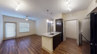 This single-floor residence offers a thoughtfully designed open-concept layout that seamlessly connects the kitchen and living area. Rich, dark-toned hardwood-style flooring runs throughout the main living spaces, lending warmth and a modern aesthetic. The kitchen serves as the centerpiece, featuring a granite-topped island with an undermount sink, sleek dark cabinetry, black appliances including a range, microwave, and refrigerator, and stylish pendant lighting overhead. A track lighting system provides additional illumination, while a ceiling fan in the living area ensures year-round comfort. The living space is bright and airy, benefiting from natural light through blinded windows and access to an exterior door. A spacious walk-in pantry or storage closet is conveniently located adjacent to the kitchen. The bedroom is generously sized, offering plush carpeting, a ceiling fan, and a large window that fills the room with natural light. The bathroom is finished to a high standard, showcasing a dual-sink granite vanity with dark cabinetry, a full-size soaking tub, large-format gray tile surround, and hardwood-style flooring. Every detail of this residence reflects a clean, contemporary design ethos with quality finishes throughout.