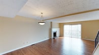 This multi-floor space showcases a welcoming living area characterized by warm hardwood flooring that extends throughout the open layout. A textured ceiling adds visual depth overhead, while a pendant light fixture provides a central focal point in the main living space. The room is anchored by a classic brick fireplace with a white-painted surround, offering a traditional design element that adds character and warmth to the area.

Natural light flows generously into the space through large sliding glass doors dressed with vertical blinds, which open onto a balcony offering views of the surrounding exterior. The neutral, warm-toned walls create a versatile backdrop that complements the rich wood tones of the flooring. A structural beam detail subtly divides the living space, adding architectural interest while maintaining an open, cohesive feel.

The overall ambiance is clean and understated, with classic finishes throughout. The combination of the fireplace, generous natural light, and balcony access gives the living area a comfortable, functional character. Spanning three floors, the property offers a substantial amount of living space across multiple levels.