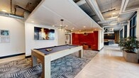 Located in the heart of Salt Lake City at 111 S 300 W, this thoughtfully designed amenity floor offers a dynamic mix of social, recreational, and relaxation spaces spread across a generously sized single-floor layout. The space blends industrial-modern architecture — exposed ductwork and concrete ceilings — with refined interior finishes throughout.

The game area anchors one end of the floor, featuring a full-size billiards table set atop geometric patterned carpet, a contemporary multi-arm chandelier overhead, and a vibrant red velvet booth nook for intimate gatherings. Adjacent to it, a well-appointed kitchen showcases white subway tile backsplash, warm wood ceiling accents, brass fixtures, stainless steel appliances, and herringbone-patterned tile flooring.

A cozy lounge area centers around a sleek linear fireplace set against a dramatic textured dark tile wall, surrounded by plush navy blue seating. Nearby, a media lounge offers large sectional sofas facing a wood-framed wall-mounted screen, with floor-to-ceiling windows and direct access to a furnished outdoor terrace with city views.

A private card room provides an intimate retreat, complete with a classic poker table, built-in bookshelves, and abundant natural light streaming through expansive windows. Together, these zones create a sophisticated and welcoming communal environment.