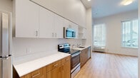 This single-floor unit offers a thoughtfully designed open layout that efficiently combines kitchen and living functions within a compact footprint. The kitchen is a standout feature, equipped with a full suite of stainless steel appliances including a refrigerator, electric range, built-in microwave, and dishwasher. Two-tone cabinetry pairs warm wood lower cabinets with crisp white upper cabinets extending toward notably tall ceilings, maximizing vertical storage space. White quartz countertops provide a clean, modern work surface throughout.

Light wood-tone flooring runs consistently throughout the entire unit, creating a sense of visual continuity and warmth. The living area benefits from multiple large windows fitted with vertical blinds, allowing generous natural light to fill the space while offering views to the exterior. A wall-mounted HVAC unit provides climate control.

The bathroom maintains the unit's modern aesthetic, featuring matching warm wood cabinetry with a white countertop vanity, a full bathtub with shower combination, chrome fixtures, and the same light wood flooring seen throughout the rest of the unit. Neutral white walls and clean lines throughout contribute to a bright, minimalist ambiance well-suited to efficient urban living.