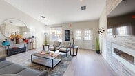 This single-floor space is designed with an open-concept layout that seamlessly connects the kitchen, dining, and lounge areas into one cohesive, functional environment. Light hardwood-style flooring runs throughout, lending warmth and visual continuity across the entire space.

The kitchen is a standout feature, anchored by an expansive island with a crisp white countertop and lined with modern bar stools, offering generous seating for casual dining or gathering. Sleek wood-toned cabinetry extends to the upper walls, complemented by stainless steel appliances and a decorative tile backsplash. Multiple windows frame outdoor views and flood the area with natural light, enhancing the airy, open feel.

Adjacent to the kitchen, a dedicated dining area accommodates a round table with upholstered seating, positioned near large windows that provide additional natural light and neighborhood views.

The lounge area offers a relaxed setting with comfortable seating arrangements, a decorative area rug, and accent furnishings. A glass-paneled door provides direct access to the outdoors, while additional windows allow natural light to permeate the room. The neutral color palette and modern décor throughout create a welcoming, contemporary ambiance well-suited for both socializing and everyday use.