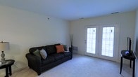 This single-floor space features a living room with a clean, understated design characterized by light-toned walls and wall-to-wall carpeting that contributes to a soft, comfortable atmosphere. The room is generously proportioned, providing ample space for seating and movement. A prominent feature is the set of white-framed French doors along one wall, which allow natural light to fill the interior while offering access to an outdoor area. The doors are fitted with multi-pane glass panels, enhancing the brightness of the room throughout the day. A floor vent is visible near the base of the wall, indicating a standard forced-air climate control system. The walls are smooth and neutral in color, offering a versatile backdrop suitable for a variety of décor styles. The ceiling is flat and uniform, maintaining a consistent, clean aesthetic throughout the room. Overall, the space presents a simple, functional living environment with a bright and airy quality driven largely by the natural light introduced through the French doors, making it a practical and adaptable space for everyday living.