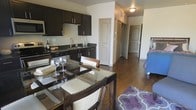 This single-floor residence showcases an open-concept layout that seamlessly integrates the kitchen, dining, and living areas into one cohesive space. The kitchen is outfitted with rich, dark espresso cabinetry paired with dark granite countertops, creating a bold, contemporary aesthetic. A stainless steel sink and built-in appliances, including a microwave mounted above the range, contribute to the kitchen's functional design. The cabinetry extends generously along the walls, offering ample storage throughout.

Adjacent to the kitchen, the dining area accommodates a glass-top table with seating for multiple guests, serving as a natural transition point between the kitchen and living space. The warm hardwood flooring runs continuously throughout, adding cohesion and a sense of warmth to the interior.

A hallway visible from the main living area leads to additional rooms, suggesting a well-organized floor plan with clearly defined private and shared zones. Neutral wall tones complement the darker finishes, keeping the space feeling bright and balanced. The overall ambiance is modern and functional, with a practical layout suited for everyday living.