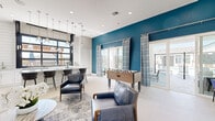 Located at Avalon Landing in Houston, TX, this single-floor community clubhouse delivers a well-appointed gathering space designed for both relaxation and socializing. The living room serves as the heart of the space, anchored by a sleek fireplace framed in textured white hexagonal tile and topped with a flat-screen TV. Plush navy leather seating, layered area rugs, and a vibrant living moss wall accent with neon signage contribute to a lively yet refined atmosphere. Recessed lighting and large black-framed glass doors flood the interior with natural light. A bold teal feature wall defines the game zone, where a foosball table and additional lounge seating create a casual entertainment hub. Pendant lighting highlights a built-in wet bar with bar-height seating, ideal for social gatherings. The space transitions seamlessly to a covered outdoor balcony outfitted with ceiling fans, round dining tables, and lounge chairs, all overlooking a resort-style pool. Shiplap ceilings and clean white exterior walls give the balcony a crisp, coastal feel. Light-toned large-format tile flooring runs throughout, unifying the entire space with a clean, modern aesthetic.
