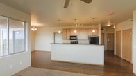 This single-floor space showcases an open-concept layout that seamlessly connects the kitchen and living areas, creating a cohesive and functional environment. The kitchen is equipped with light maple cabinetry offering generous storage, dark granite countertops, and black appliances including a built-in microwave and refrigerator. A raised breakfast bar serves as a natural divider between the kitchen and the adjoining living space, providing both a casual dining option and a visual anchor for the room. Three pendant lights hang above the kitchen area, complementing a track lighting fixture that provides focused illumination over the workspace. The living area features warm hardwood flooring that transitions to carpet, adding textural variety throughout the space. A ceiling fan positioned centrally aids in air circulation, while a chandelier near the dining area introduces an elegant touch. The neutral wall tones and warm cabinetry contribute to an inviting ambiance. The layout is well-suited for everyday living, offering clear sightlines and an efficient flow between the kitchen, dining, and living zones.