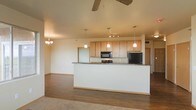 This single-floor space showcases an open-concept layout that seamlessly connects the kitchen and living areas, creating a cohesive and functional environment. The kitchen is equipped with light maple cabinetry offering generous storage, dark granite countertops, and black appliances including a built-in microwave and refrigerator. A raised breakfast bar serves as a natural divider between the kitchen and the adjoining living space, providing both a casual dining option and a visual anchor for the room. Three pendant lights hang above the kitchen area, complementing a track lighting fixture that provides focused illumination over the workspace. The living area features warm hardwood flooring that transitions to carpet, adding textural variety throughout the space. A ceiling fan positioned centrally aids in air circulation, while a chandelier near the dining area introduces an elegant touch. The neutral wall tones and warm cabinetry contribute to an inviting ambiance. The layout is well-suited for everyday living, offering clear sightlines and an efficient flow between the kitchen, dining, and living zones.