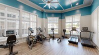 Located in North Charleston, SC, this fitness center offers a well-appointed exercise space designed to support a comprehensive workout routine. The room is distinguished by its striking teal upper walls contrasted with crisp white wainscoting panels, creating a vibrant and energizing atmosphere. Large multi-pane windows along multiple walls flood the space with natural light and provide views of the surrounding exterior, enhancing the open, airy feel. A coffered ceiling with a ceiling fan and recessed lighting adds architectural character overhead.

The durable hardwood-style flooring provides a clean, resilient surface throughout. Cardio equipment is well represented, including treadmills, ellipticals, stair climbers, stationary bikes, and a rowing machine. A wall-mounted flat-screen TV faces the cardio area, making workouts more engaging. Functional training tools such as a vertical knee raise station, medicine ball rack, yoga mats, foam rollers, and a BOSU balance trainer round out the offerings. The layout is thoughtfully organized to maximize usability while maintaining clear, open floor space for stretching and floor exercises. Multiple HVAC vents ensure comfortable climate control year-round, making this fitness center a practical and inviting destination for daily exercise.