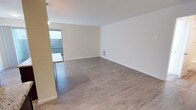 This single-floor unit offers a clean, contemporary design throughout. The open-concept living area is bright and airy, featuring light-toned laminate wood flooring and neutral white walls that maximize the sense of space. A sliding glass door leads to a private balcony enclosed by a slatted privacy screen, allowing natural light to fill the room.

The kitchen is a standout feature, showcasing rich dark mahogany cabinetry paired with light granite countertops for a striking contrast. Stainless steel appliances — including a range, microwave, refrigerator, and dishwasher — add a modern touch. A peninsula with a pendant light overhead provides additional counter space and serves as a natural divider between the kitchen and living areas.

The bedroom is generously proportioned with the same laminate flooring and crisp white walls. A sliding double-door closet offers practical storage, while a window brings in ample natural light. An adjoining full bathroom features a clean, all-white tiled tub-shower combination with chrome fixtures, maintaining the unit's consistent, modern aesthetic.

Overall, this space presents a cohesive, updated interior with functional design elements, making it an informative and practical living environment suited for everyday comfort.