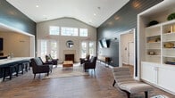 Located at 3973 Woolcock Dr in Myrtle Beach, SC, this single-story community clubhouse combines coastal charm with contemporary design across a thoughtfully arranged floor plan. The centerpiece of the space is a dramatic lounge area distinguished by soaring vaulted ceilings, a striking dark shiplap accent wall, and a statement fireplace flanked by large windows and glass French doors that flood the room with natural light. Comfortable seating arrangements, rich wood-look flooring, and a layered area rug contribute to a warm, welcoming ambiance. A built-in white shelving unit with curated décor adds a refined, library-inspired touch to one corner of the lounge. Adjacent to the main gathering area is a leasing or reception office, featuring hardwood-style flooring, natural window light, and direct sightlines to what appears to be a fitness facility with glass doors and dark shiplap detailing. A dedicated business center offers individual workstations with computers, bright window lighting, and a quiet, productive atmosphere. The fully equipped community kitchen is a standout feature, boasting deep navy cabinetry, a quartz waterfall island with bar seating, stainless steel appliances, and a subway tile backsplash — ideal for gatherings and events. Throughout, the design maintains a cohesive modern-coastal aesthetic.